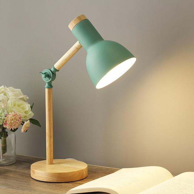 Green and wooden desk lamp on a table with a book and flowers in the background.